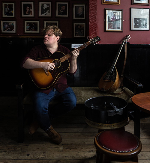 Caught mid-song, Macdara's face stretches with intensity as he plays his guitar in a darkened room in front of wall of interior glass.